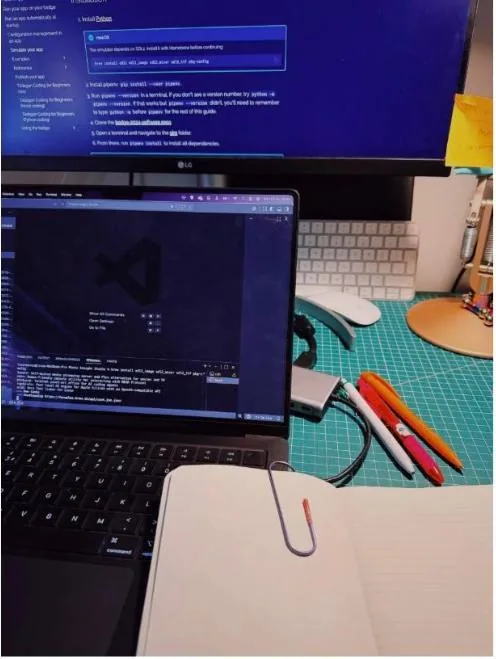 A laptop running VS Code beside a cutting mat, notebook and plotter pens